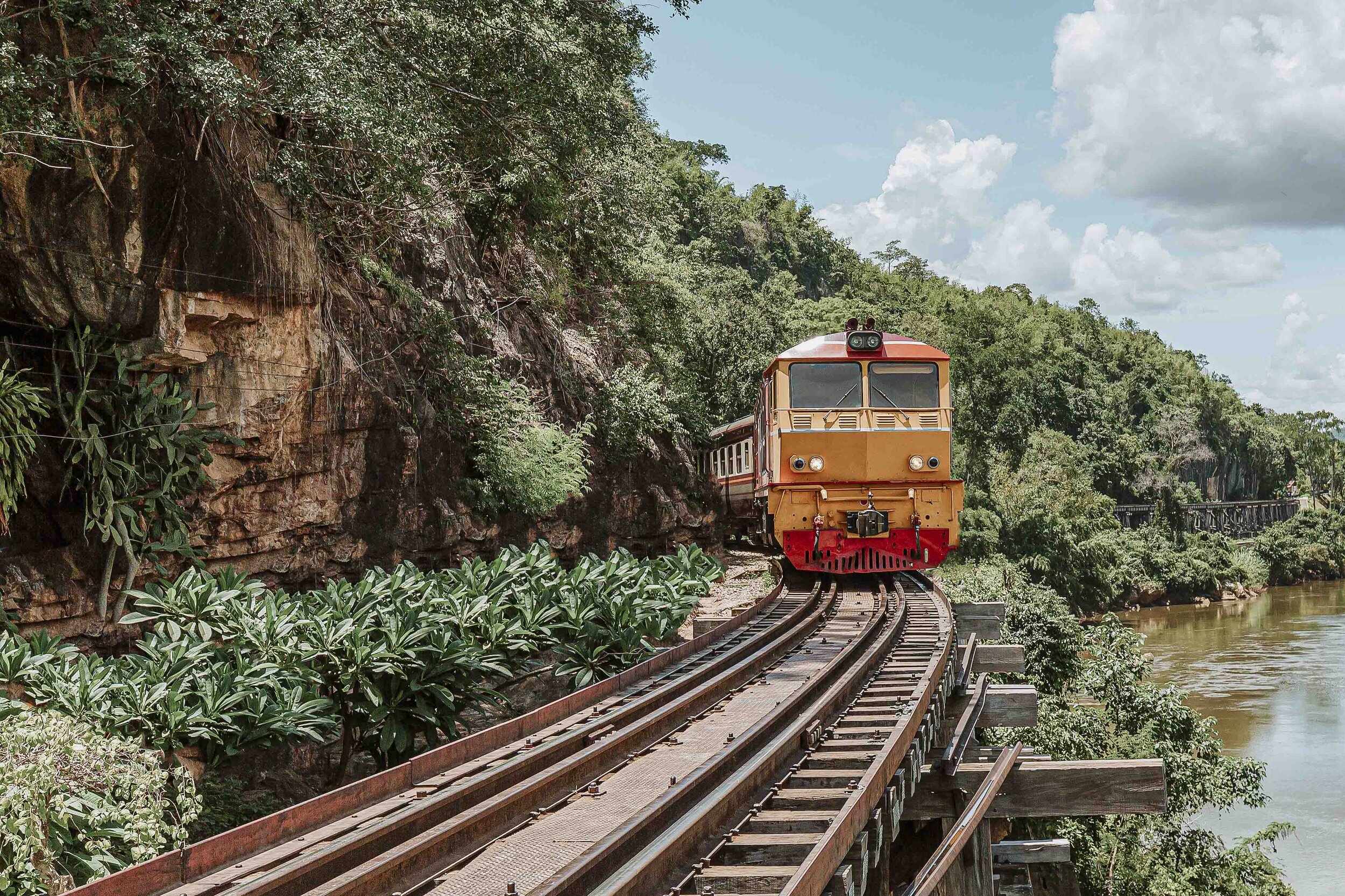 A yellow train passing on the tracks by the river in Kanchanaburi - Things to do on your 7 days in thailand itinerary
